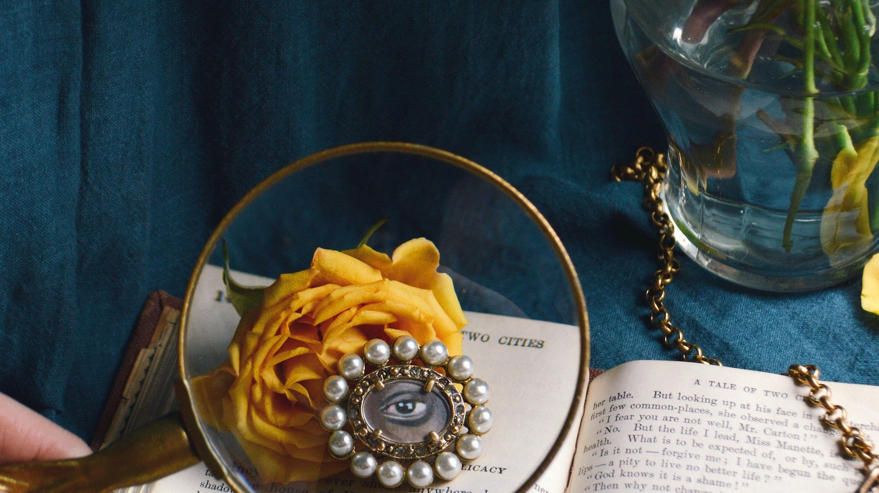 Against a dramatic, deep blue background, golden roses rest in a glass vase and on top of an antique book open to display French text. A white person's hand holds a magnifying glass over the rose and book, and at its center is the enlarged image of a hori