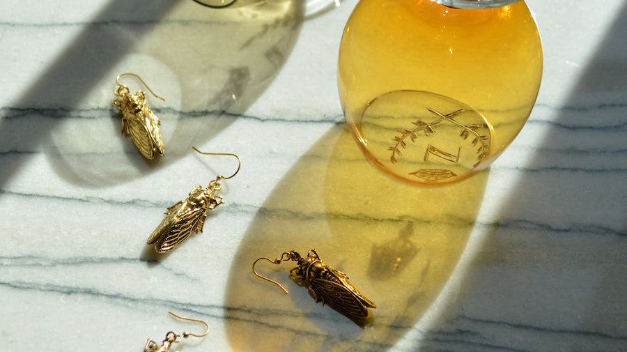 A scattering of four, golden cicadas with detailed wings and heads rests over dark-veined white marble, two amber-colored beer glasses perched above with sunlight streaming through.