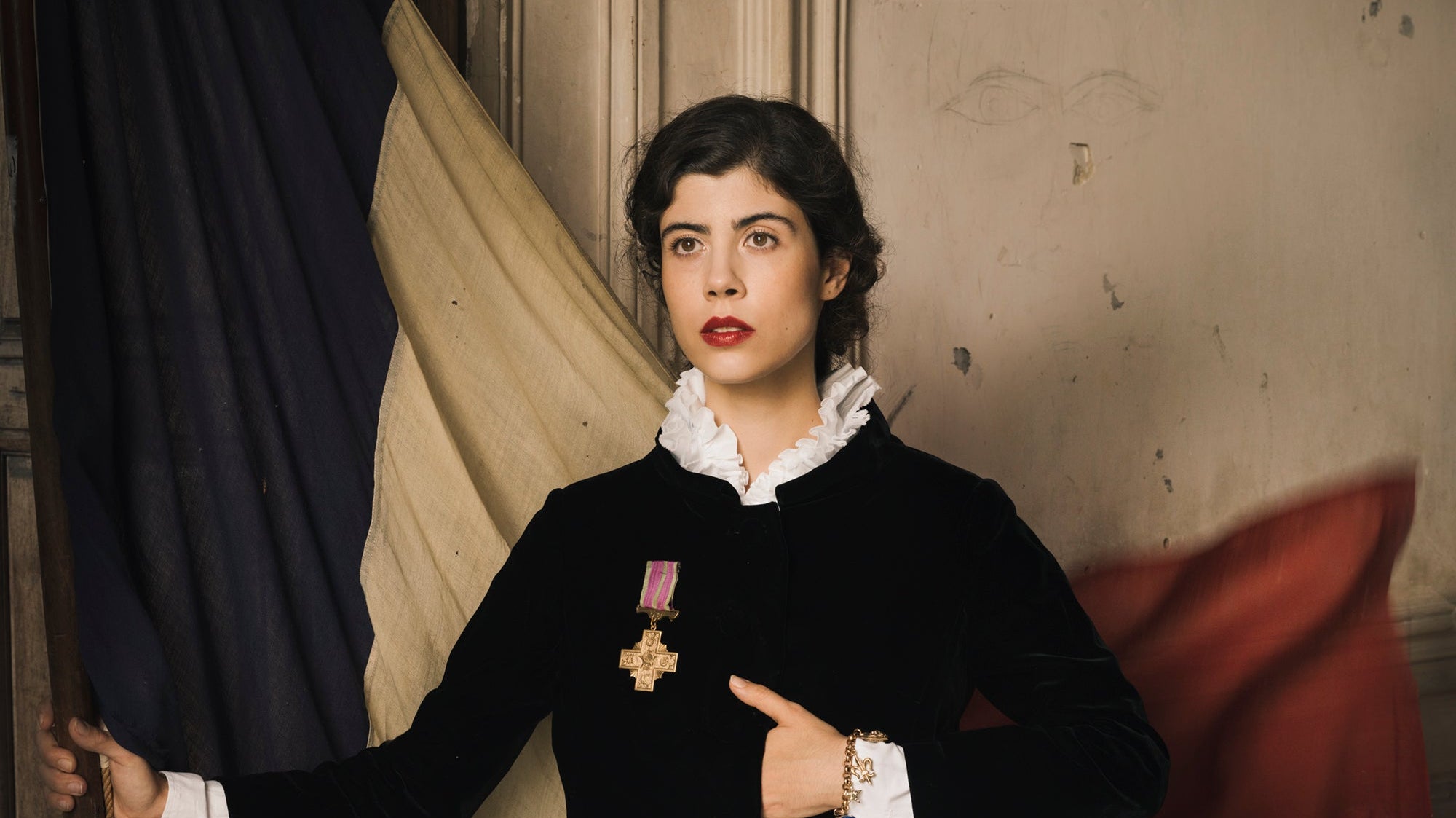 A powerful image of a white Frenchwoman in a black vintage-feeling jacket stands holding a massive, antique French flag with her hand tucked beneath her breast pocket of her jacket.