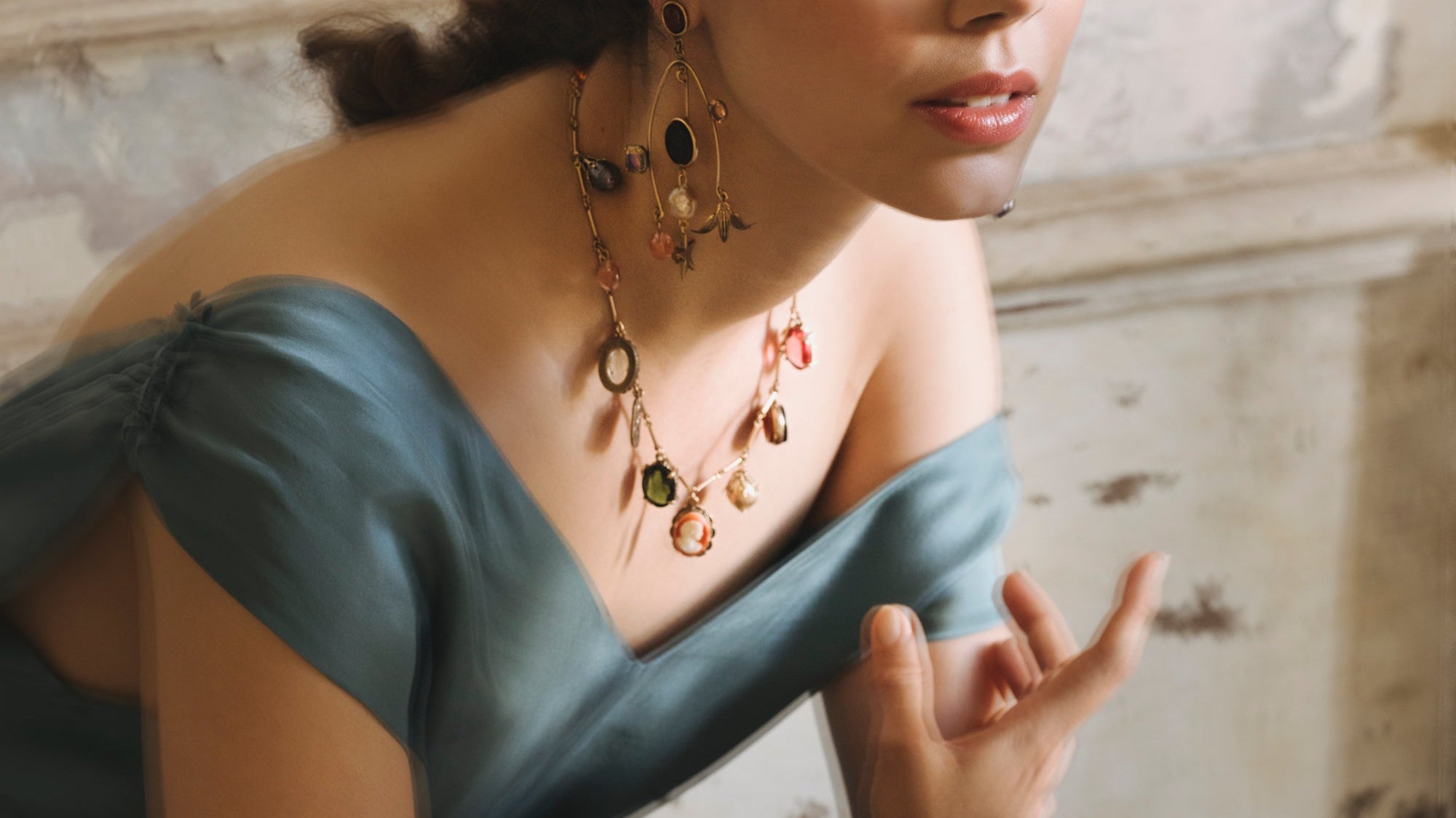 A romantic image of a white woman with dark brown, curly hair and round, warm brown eyes, she leans over in a vintage blue silk dress, one hand extended before her as if in conversation. Around her wrist, throat, and dangling from either ear hang charms.