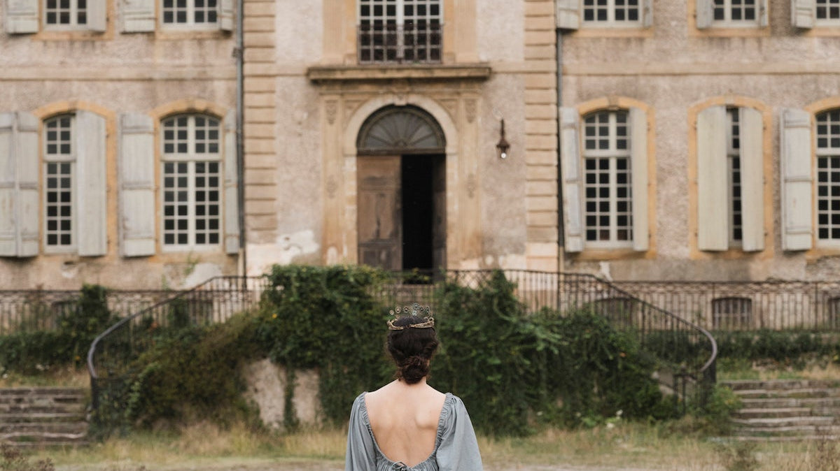 A white woman wearing an old, light blue dress walks toward a dusty blue and tan French chateau down a dust-strewn path in an old world romantic image similar to a painting.