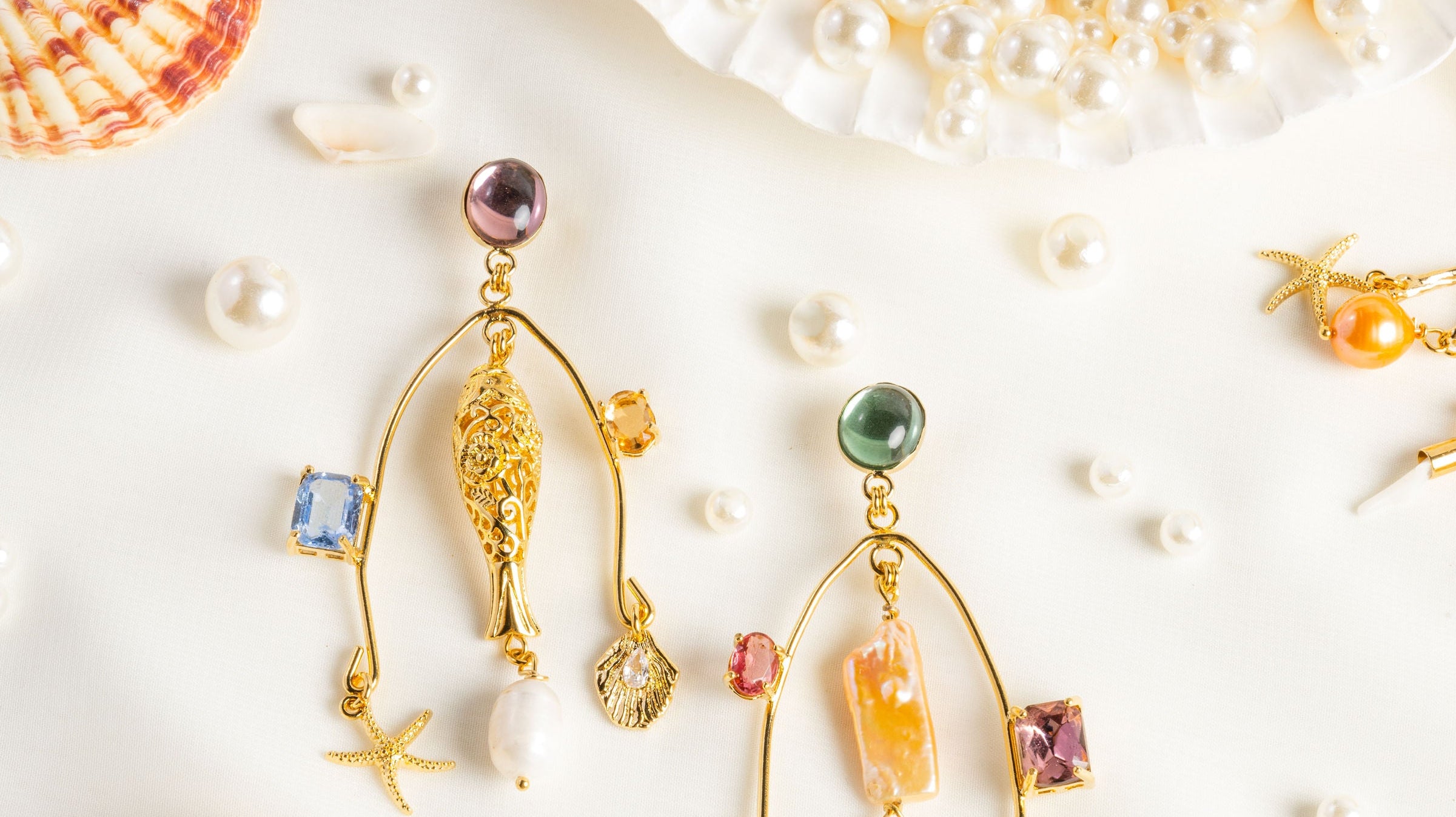 A pair of gold chandelier earrings with fish sea charms and colors lays over a white background surrounded by pearls and shells.