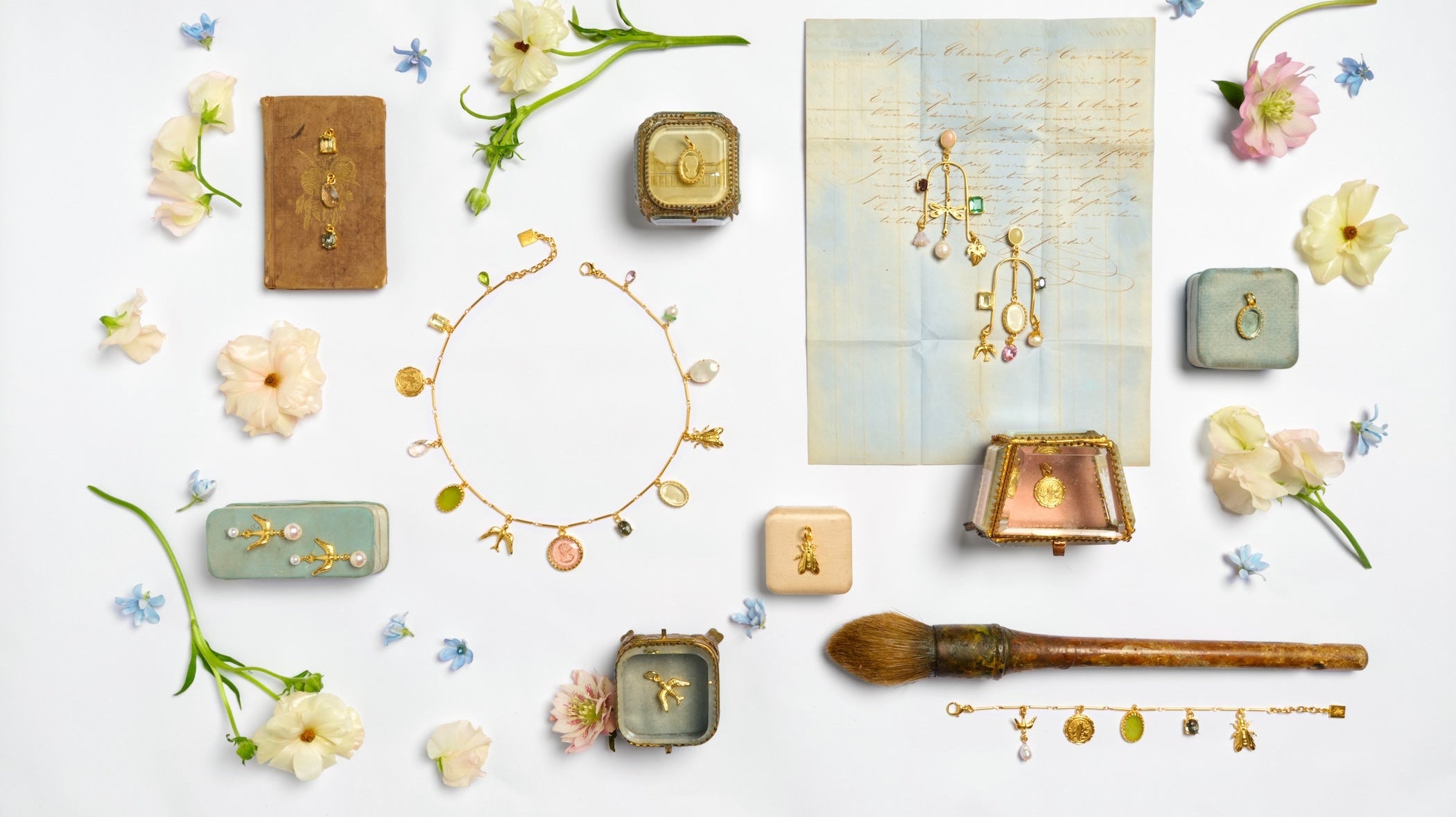 A charm necklace full of gold and German glass charms in a circle in the middle  surrounded by jewelry boxes and fresh flowers