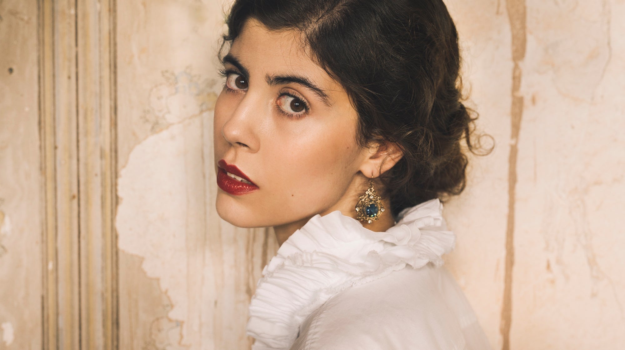 A powerful image of a white Frenchwoman wearing an antique white collared shirt against an antique mural. She looks directly at the camera with deep brown eyes, a regal blue-stoned earring hanging below her lobe.