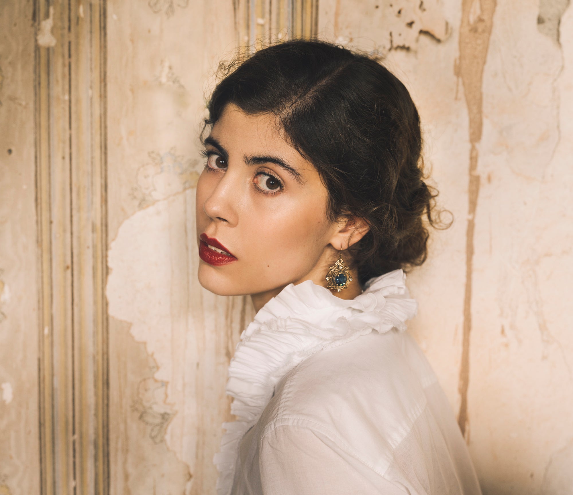 A powerful image of a white Frenchwoman wearing an antique white collared shirt against an antique mural. She looks directly at the camera with deep brown eyes, a regal blue-stoned earring hanging below her lobe.