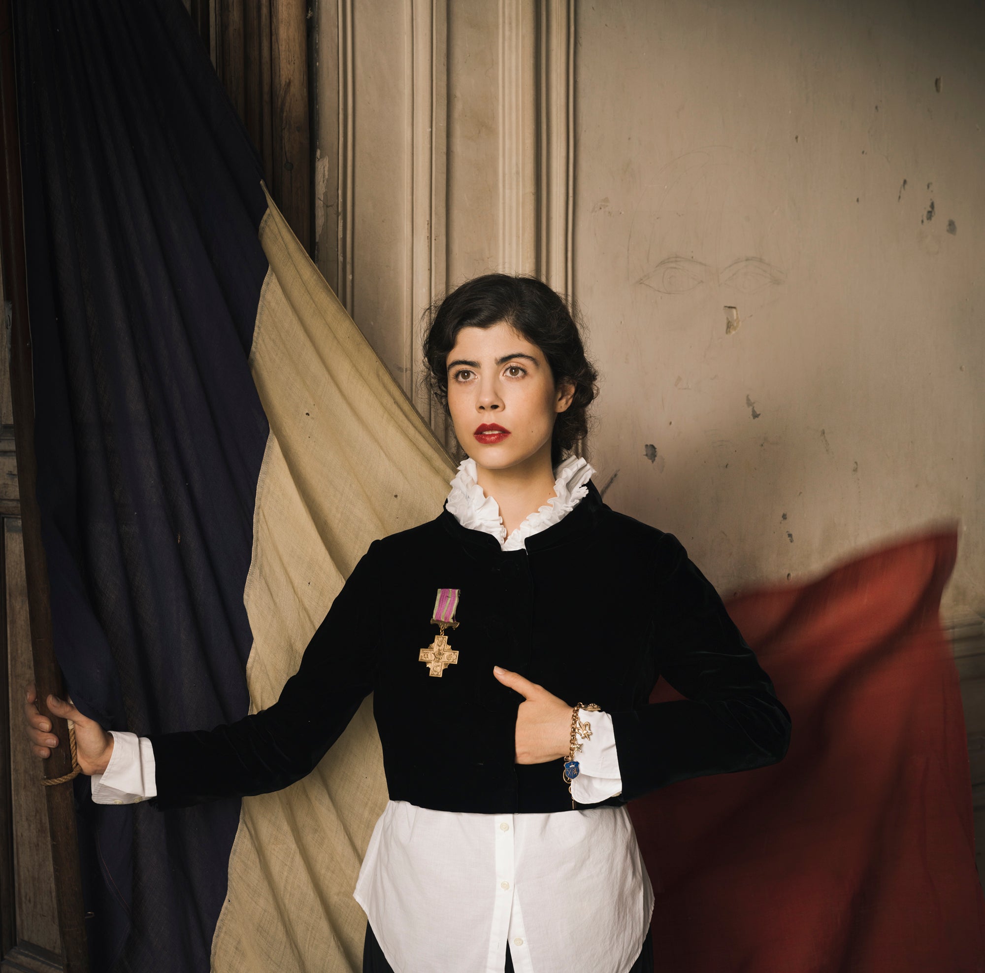 A powerful image of a white Frenchwoman in a black vintage-feeling jacket stands holding a massive, antique French flag with her hand tucked beneath her breast pocket of her jacket.