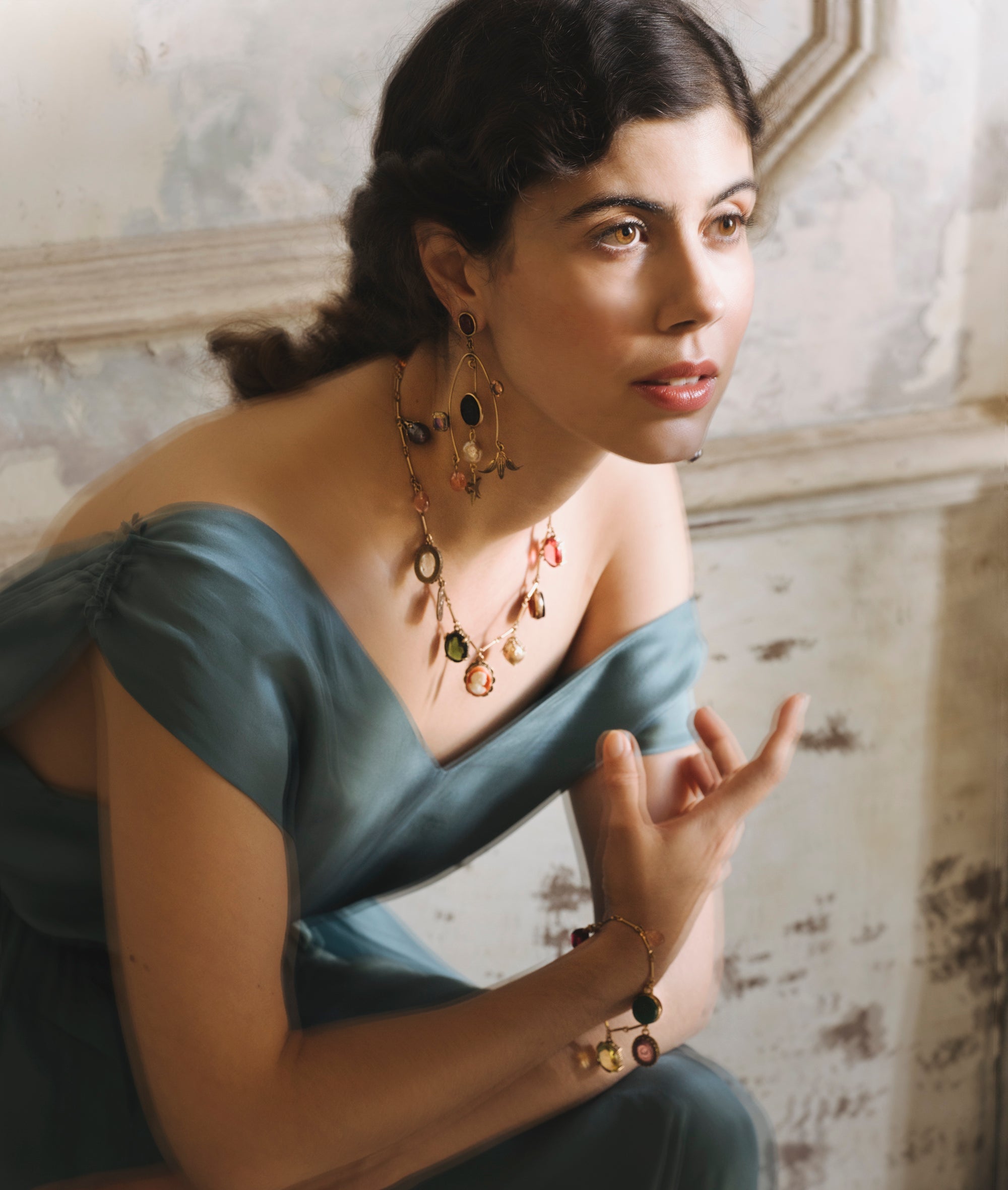 A romantic image of a white woman with dark brown, curly hair and round, warm brown eyes, she leans over in a vintage blue silk dress, one hand extended before her as if in conversation. Around her wrist, throat, and dangling from either ear hang charms.