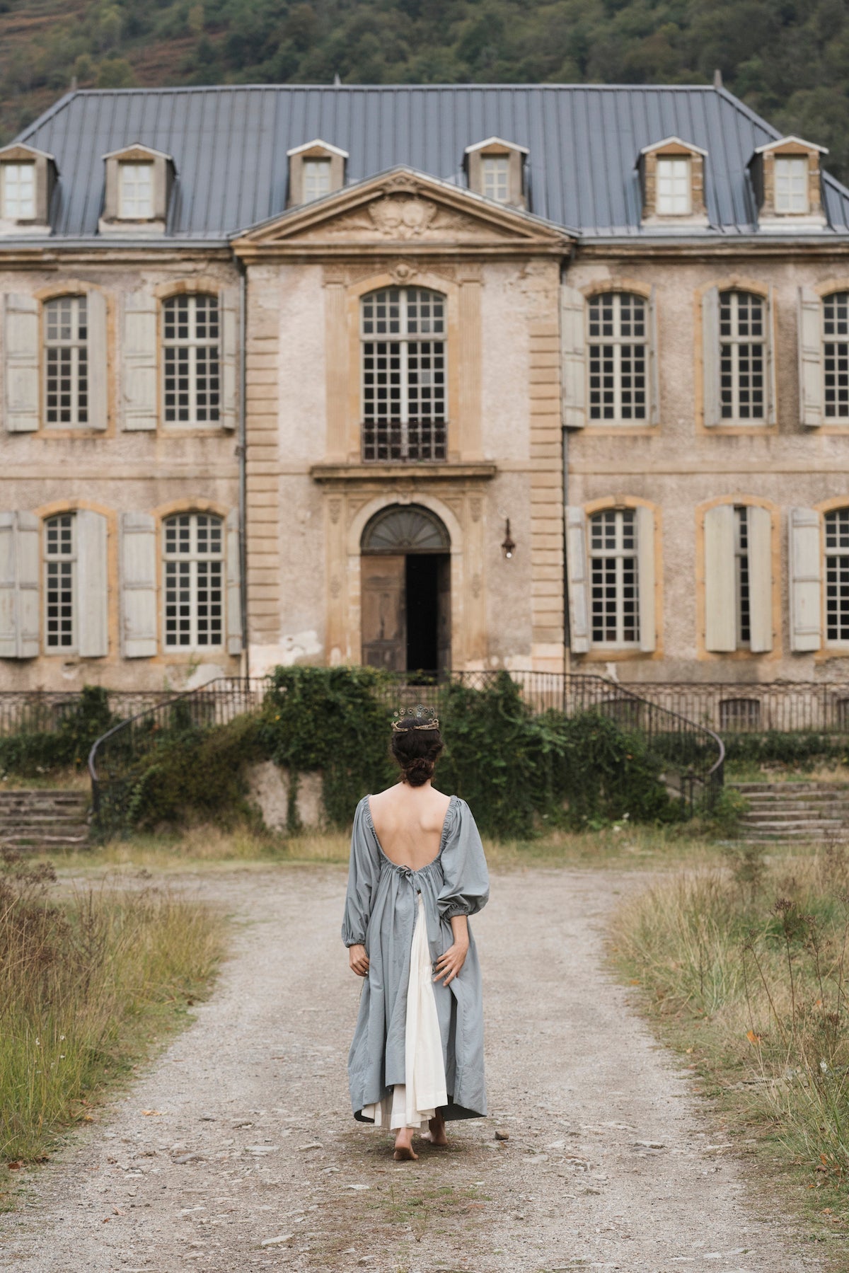 A white woman wearing an old, light blue dress walks toward a dusty blue and tan French chateau down a dust-strewn path in an old world romantic image similar to a painting.