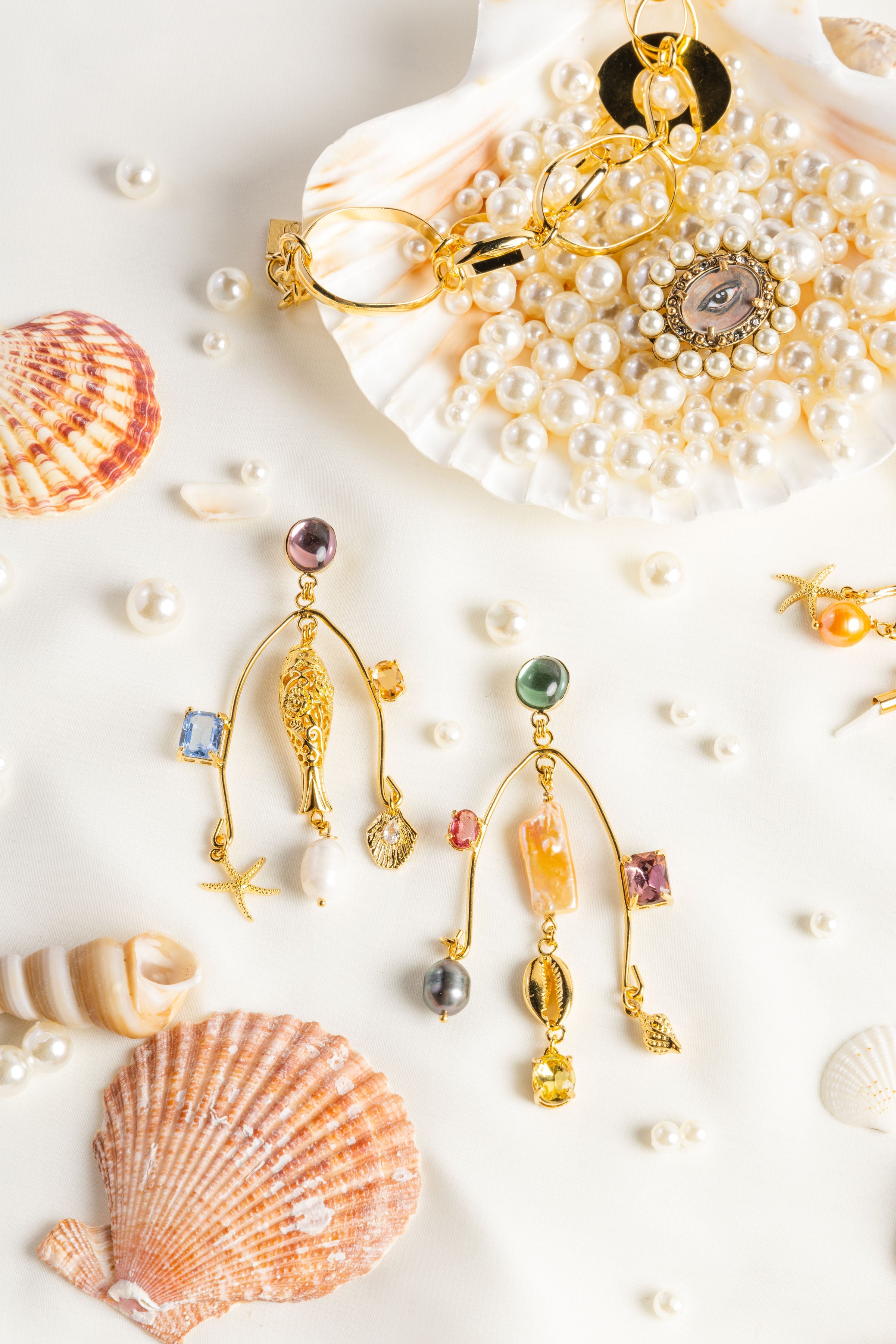 A pair of gold chandelier earrings with fish sea charms and colors lays over a white background surrounded by pearls and shells. 