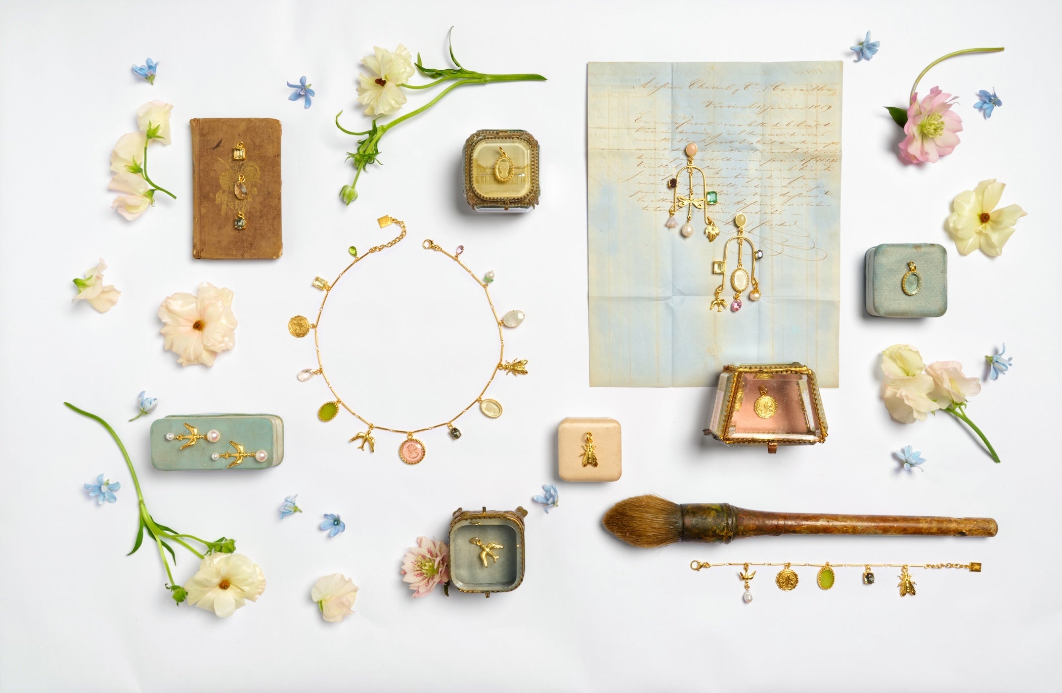 A charm necklace full of gold and German glass charms in a circle in the middle  surrounded by jewelry boxes and fresh flowers
